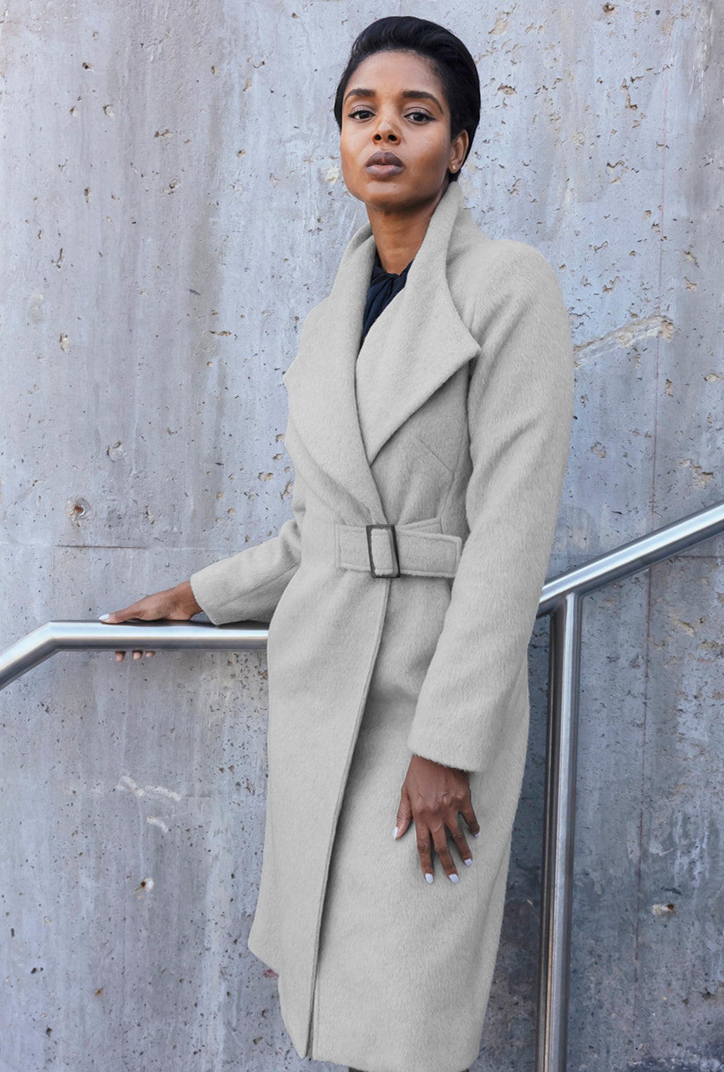 Woman wearing a long beige brushed wool coat standing against a concrete wall.